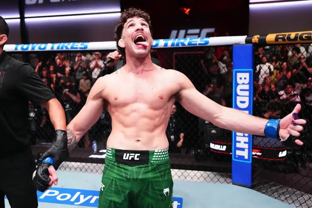 UFC fighter shirtless in green shorts raises arms in triumph inside the octagon, screaming with crowd cheering behind him.