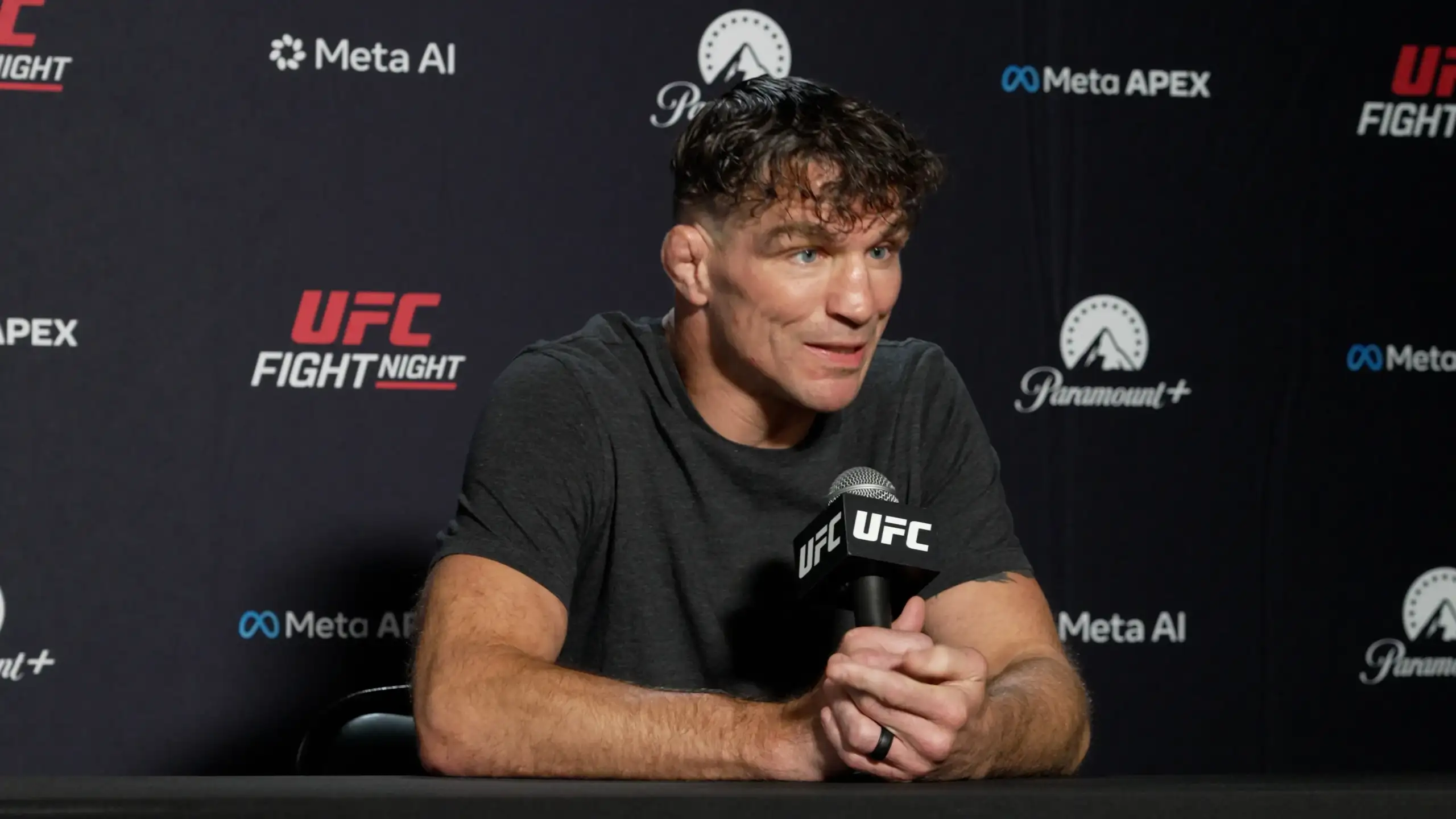 Man at a UFC press conference speaking into a UFC microphone, backdrop shows sponsor logos like Meta AI and Paramount+.