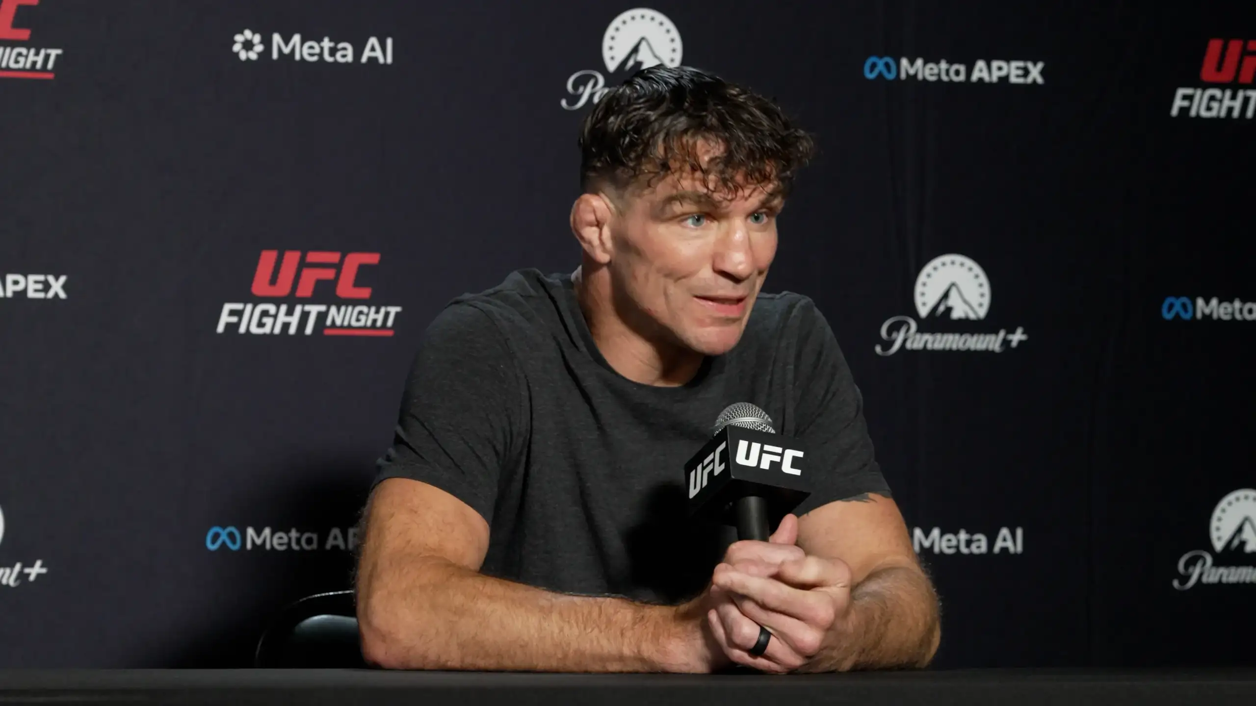 MMA fighter at a UFC press conference speaking into a handheld UFC microphone with a dark backdrop and sponsor logos behind him.