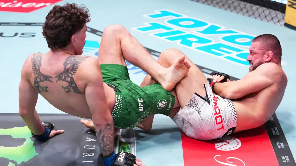 Two male mixed martial artists grapple on the mat inside a cage, one in green shorts and the other in white shorts, their legs tangled in a clinch near the canvas logos.