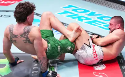 Two male mixed martial artists grapple on the mat inside a cage, one in green shorts and the other in white shorts, their legs tangled in a clinch near the canvas logos.