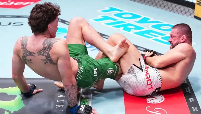 Two male mixed martial artists grapple on the mat inside a cage, one in green shorts and the other in white shorts, their legs tangled in a clinch near the canvas logos.