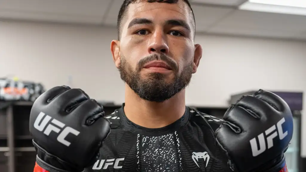 Male mixed martial artist wearing UFC gloves and a black rash guard, facing the camera in a gym.”