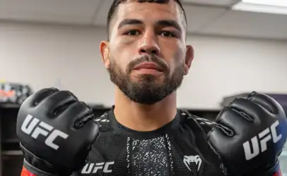 Male mixed martial artist wearing UFC gloves and a black rash guard, facing the camera in a gym.”