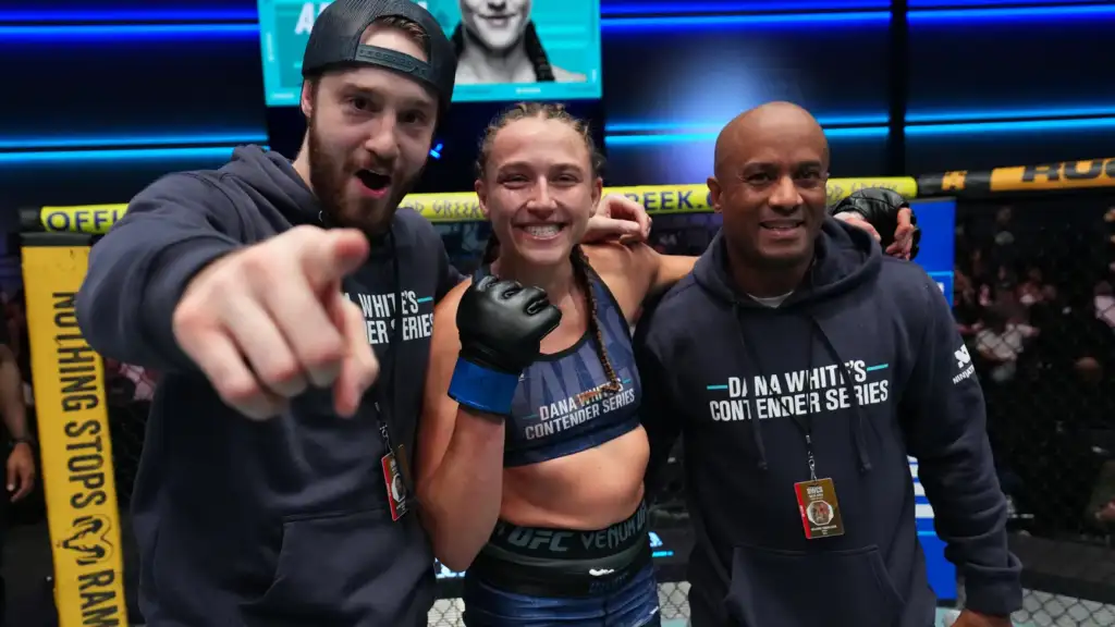 Three people inside a UFC cage: a smiling female fighter with braided hair and blue gloves center, flanked by two coaches in matching hoodies.