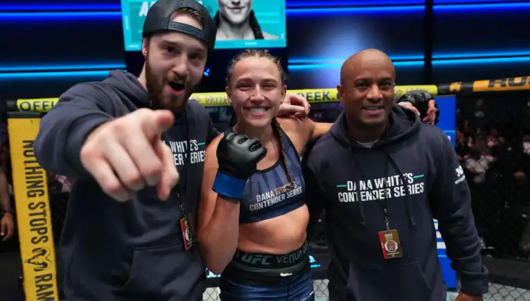 Three people inside a UFC cage: a smiling female fighter with braided hair and blue gloves center, flanked by two coaches in matching hoodies.
