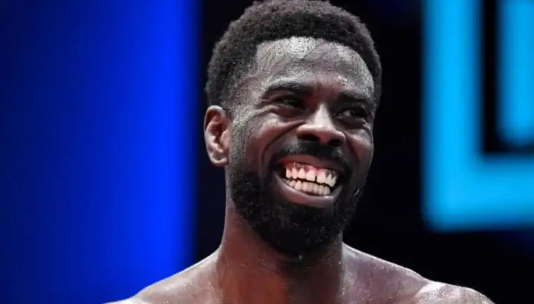 Close-up of a smiling man with a beard, sweat on his face, on a stage with blue lighting in the background