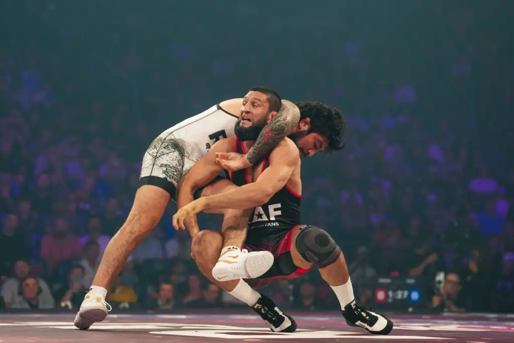 Two male wrestlers in a high‑flying hold during a match, one with a tattooed arm wearing white, the other in black and red.