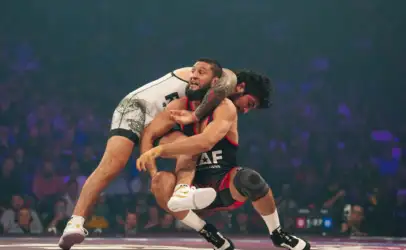 Two male wrestlers in a high‑flying hold during a match, one with a tattooed arm wearing white, the other in black and red.