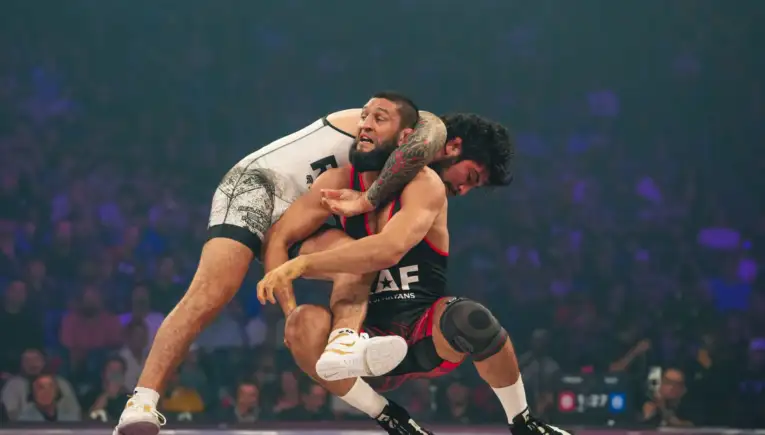 Two male wrestlers in a high‑flying hold during a match, one with a tattooed arm wearing white, the other in black and red.
