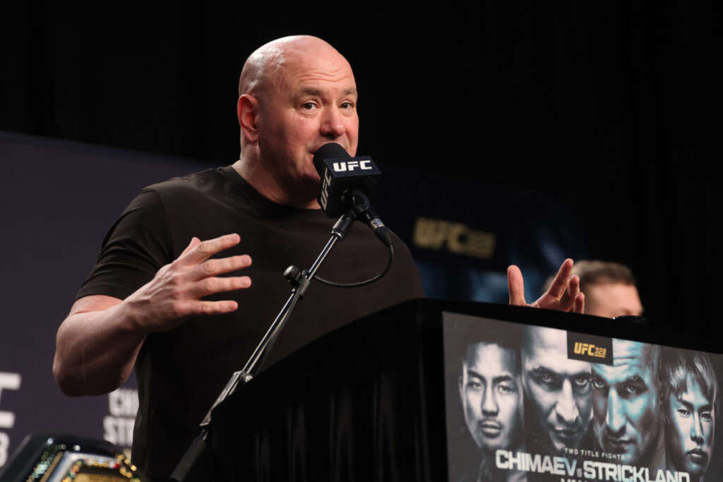 Man in a black shirt speaks into a UFC microphone at a podium, with a UFC 328 poster featuring Chimaev and Strickland in the background.