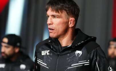 Male UFC fighter in a black zip-up jacket, looking to the side and speaking or listening backstage.