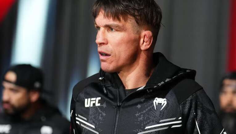 Male UFC fighter in a black zip-up jacket, looking to the side and speaking or listening backstage.