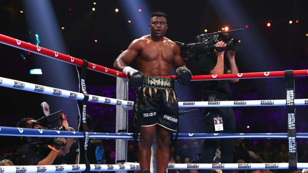 Boxer stands in the ring shirtless, wearing black and gold shorts, surrounded by cameras and spotlights upstage.