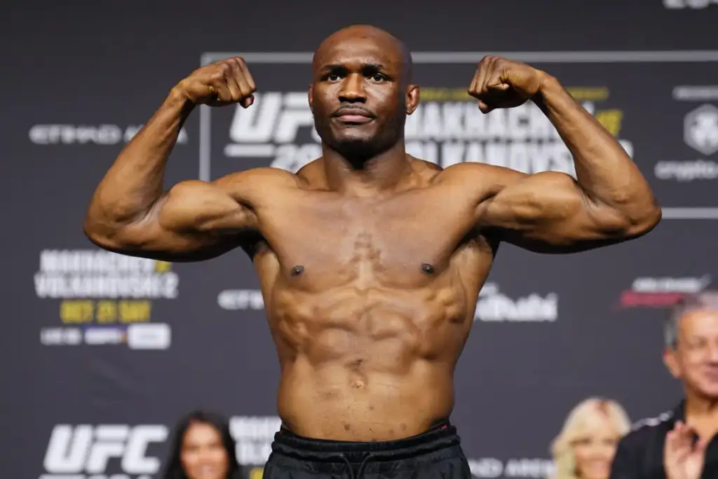 Male professional bodybuilder flexing biceps on a weigh-in stage, shirtless, with UFC banners in the background.