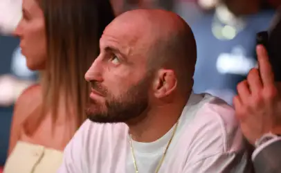 Close-up of a bald man with a beard looking to the left at a social event; wearing a white shirt and gold chain.