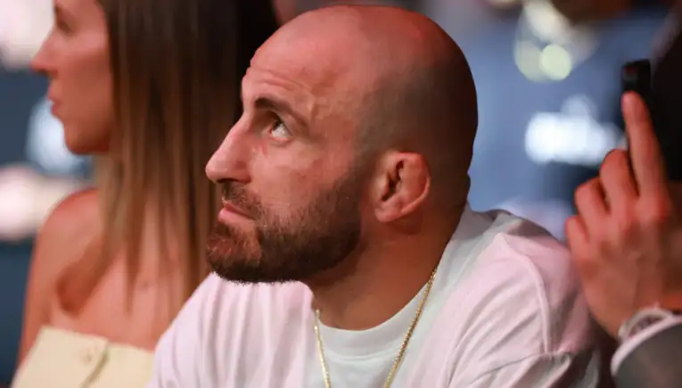 Close-up of a bald man with a beard looking to the left at a social event; wearing a white shirt and gold chain.