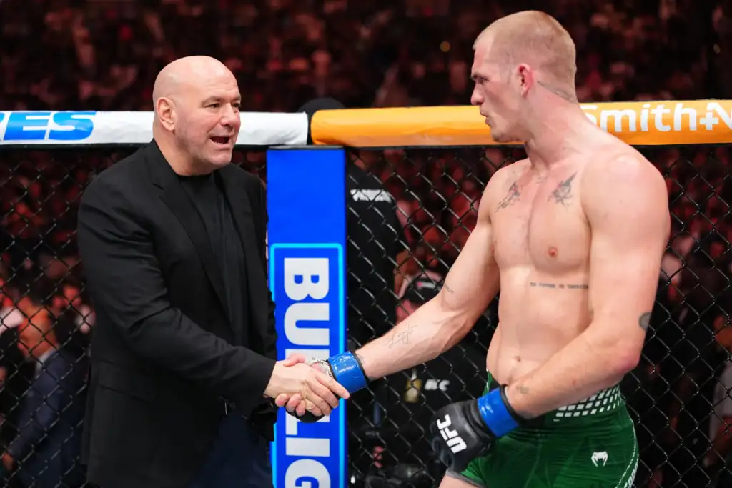 Bald man in a black suit shakes hands with a shirtless MMA fighter inside a fence-lined cage.