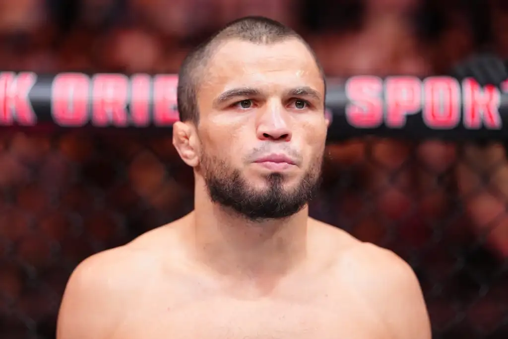 Close-up portrait of a shirtless male mixed martial artist with a trimmed beard, inside a cage with red signage in the background.