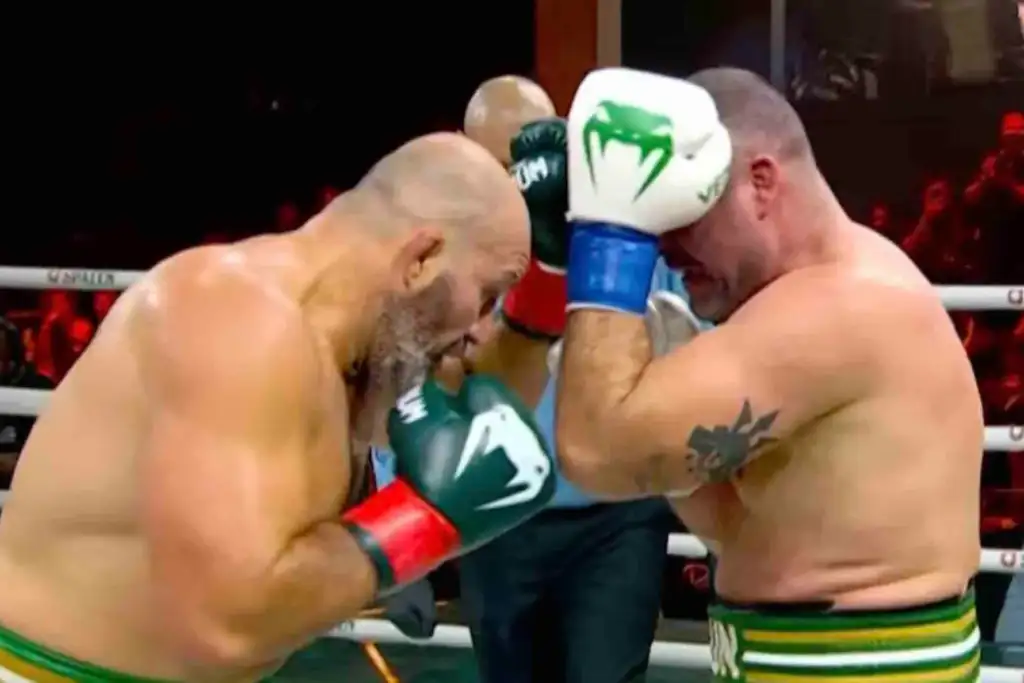 Two muscular boxers lean in with foreheads touching during a bout, wearing green-trimmed trunks and gloves as the referee watches in the background.