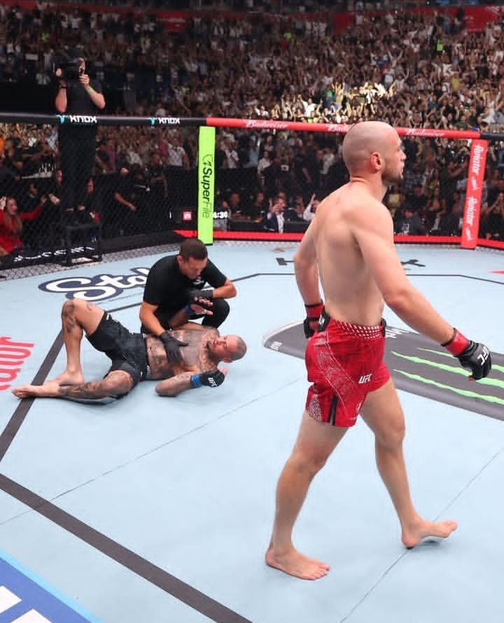 UFC octagon scene: a bald fighter in red shorts walks away as an opponent on the mat is checked by a referee amid a cheering crowd.