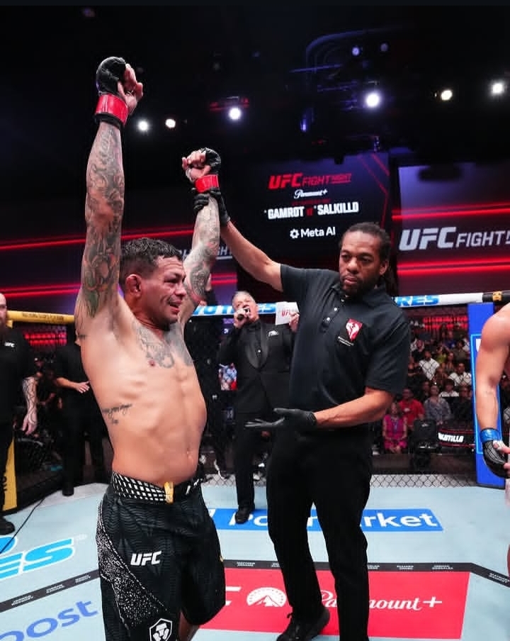 UFC fighter raises both arms in victory inside the octagon as referee signals win, crowd watching in the background.