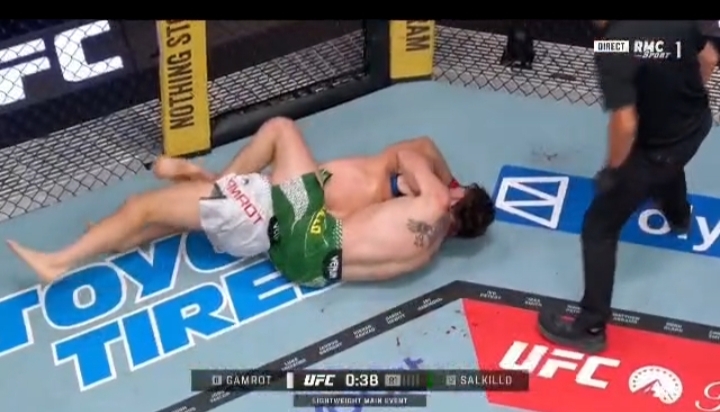 Two male mixed martial artists grapple on the mat inside a UFC Octagon while a referee stands nearby. One fighter wears green shorts and is on top in a clinched position.