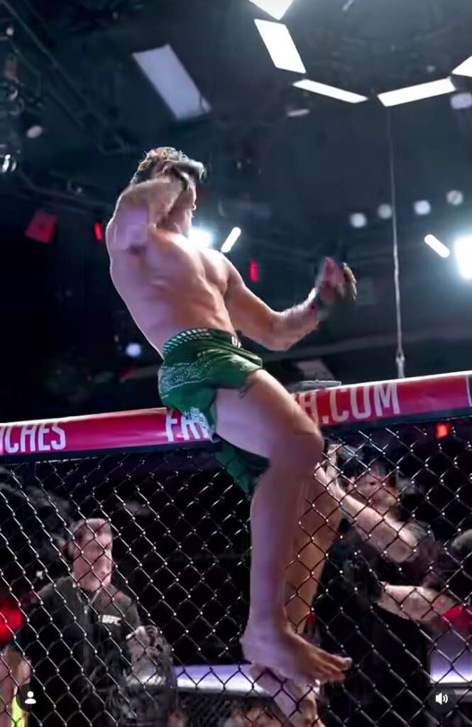 Muscular male fighter climbs the cage wall during a mixed martial arts match, wearing green shorts and no shirt.