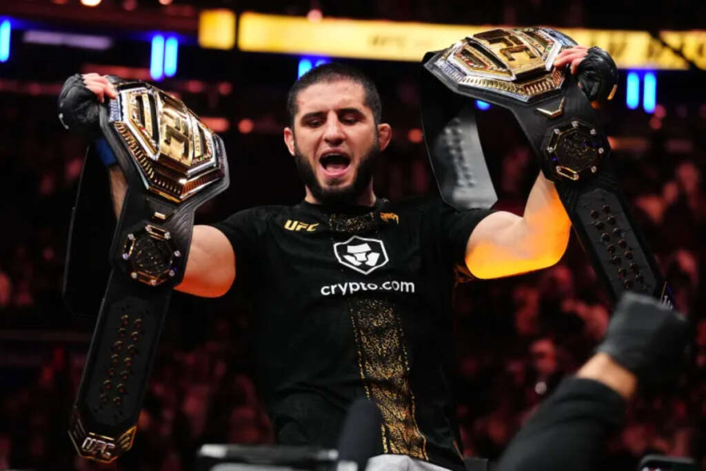 Islam-Makhachev-will-not-retire-following-his-UFC-330-title-defense-against-Ian-Machado-Garry - AfroCombat Male UFC fighter celebrates with two championship belts raised high, crowd in background, wearing a black shirt with crypto.com logo.