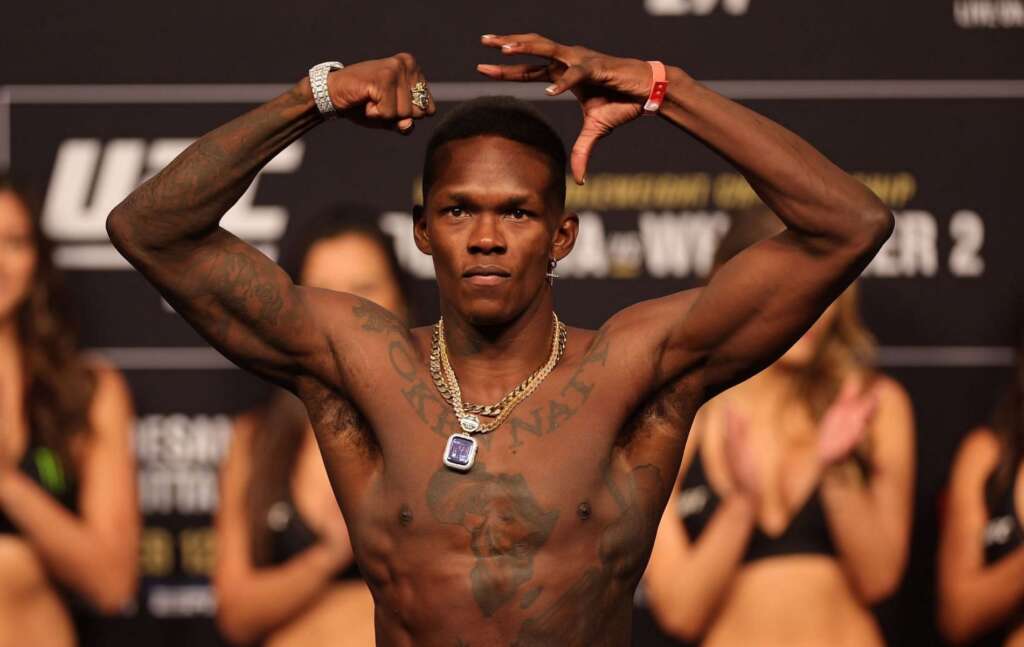Shirtless African American fighter with arms raised, showing tattoos and chain necklaces at a weigh-in stage with blurred audience behind him.