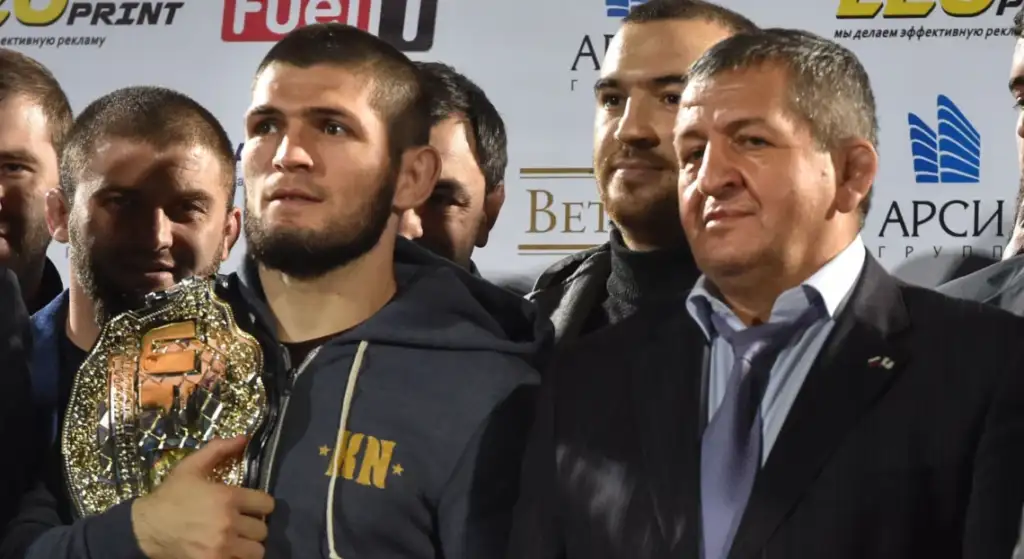 Group of men posing for a photo at a sports awards event; a bearded man in a gray hoodie holds a shiny championship belt in front of logos.