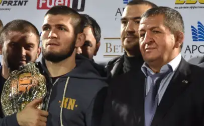 Group of men posing for a photo at a sports awards event; a bearded man in a gray hoodie holds a shiny championship belt in front of logos.