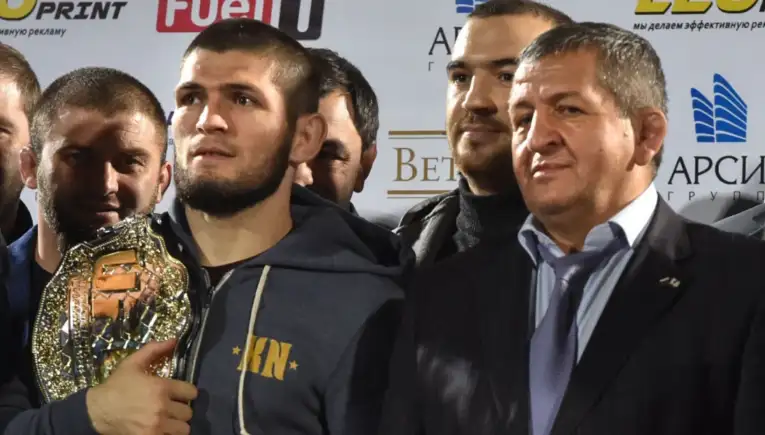 Group of men posing for a photo at a sports awards event; a bearded man in a gray hoodie holds a shiny championship belt in front of logos.