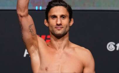 Smiling shirtless man on stage raises his arm in victory, tattoos visible on his left arm.