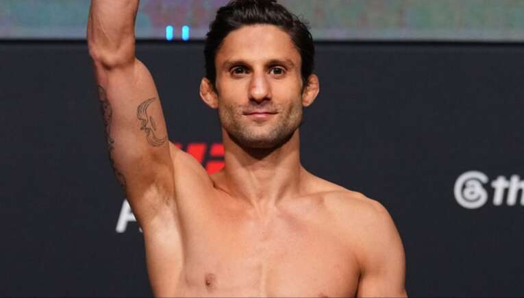 Smiling shirtless man on stage raises his arm in victory, tattoos visible on his left arm.
