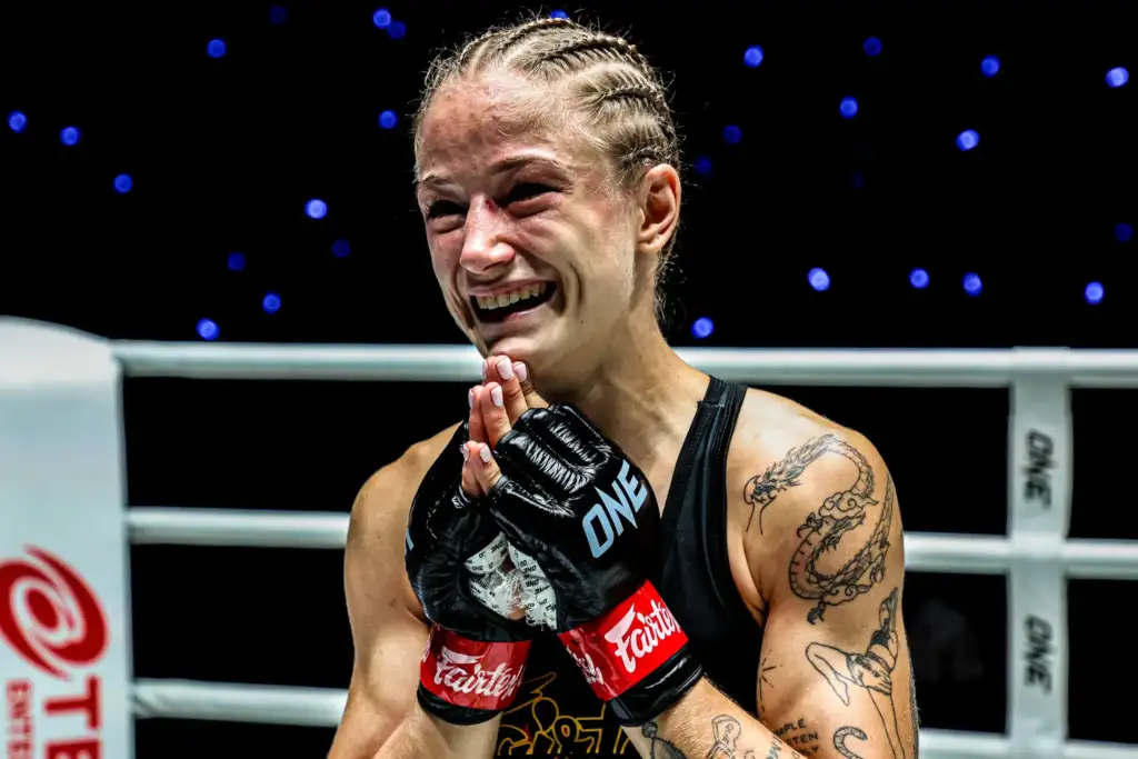 Female mixed martial artist smiling with gloved hands pressed together in a post-fight gesture inside the ring, tattoos visible on her arm.