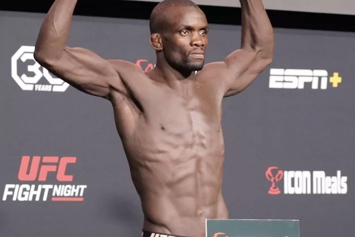 Male UFC fighter stands shirtless on a weigh‑in stage, arms raised in victory, with ESPN+ and UFC Fight Night logos in the background.