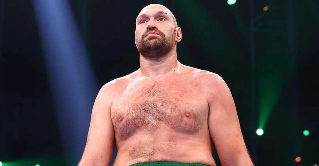 Shirtless muscular man with a beard standing in a dim arena, looking upward under blue and green stage lights.