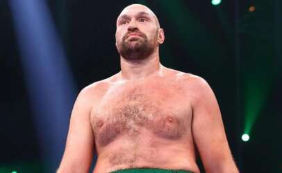 Shirtless muscular man with a beard standing in a dim arena, looking upward under blue and green stage lights.