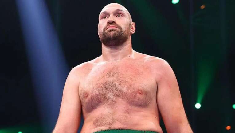 Shirtless muscular man with a beard standing in a dim arena, looking upward under blue and green stage lights.