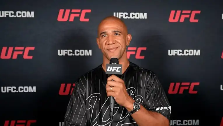 Man in a black striped shirt speaks into a UFC microphone against a UFC backdrop.