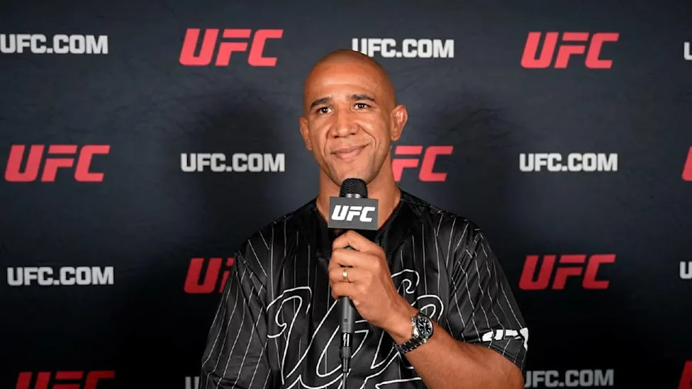 Man in a black striped shirt speaks into a UFC microphone against a UFC backdrop.