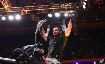 Male fighter lifts two championship belts in victory, cameras filming as crowd cheers in the arena.