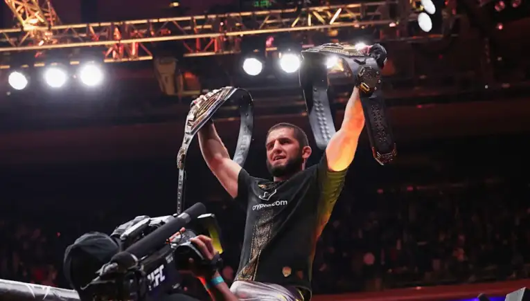 Male fighter lifts two championship belts in victory, cameras filming as crowd cheers in the arena.