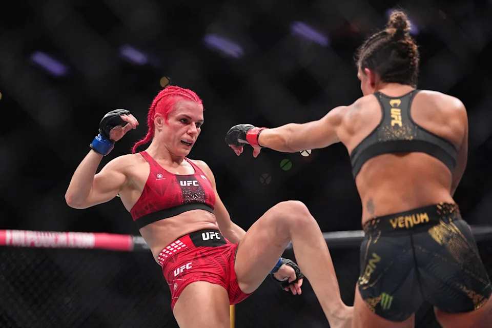 Two female UFC fighters in a cage exchange kicks mid-fight, one in red shorts delivering a kick toward the opponent in black and gold gear.