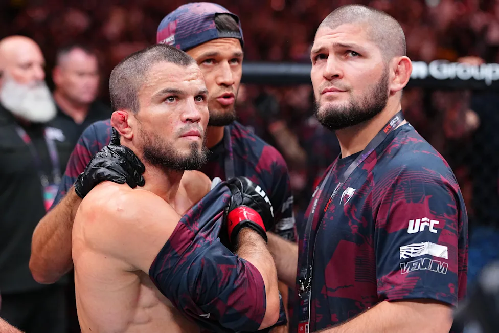 Two UFC fighters embrace in the octagon, one shirtless with red gloves and a determined expression, a teammate behind them.