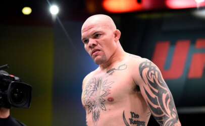 Shirtless, bald tattooed fighter with a large chest and arm tattoo, standing in a UFC arena with a camera in the foreground.