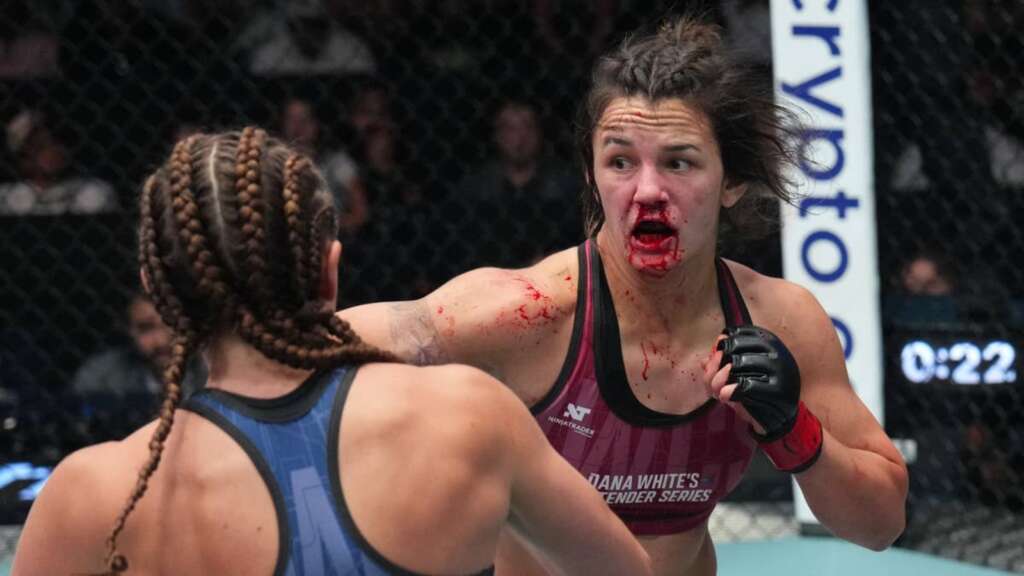 Female mixed martial artist with blood on her face lunges at her opponent inside a cage during a match.