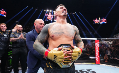 UFC fighter with a championship belt stands in the octagon, smiling as teammates applaud behind him and a crowd cheers.