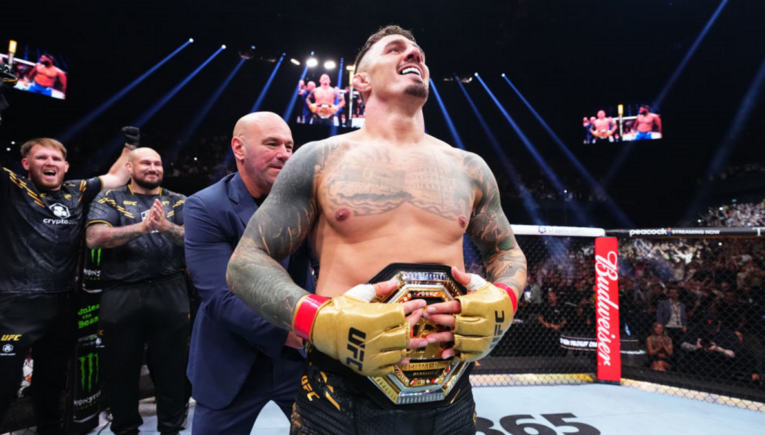 UFC fighter with a championship belt stands in the octagon, smiling as teammates applaud behind him and a crowd cheers.